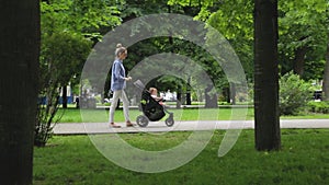 Cute slim mother with stroller walks with her infant daughter among green trees in pictorial city park