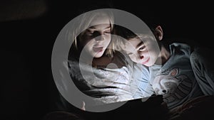 Cute small boy and girl lying under the blanket at night and watching video on tablet computer.