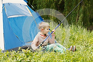 Cute small boy sits near a tent with a fishing rod in his hands.