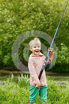 Cute small boy stand near a river with a fishing rod in his hands.