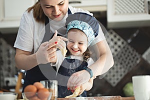 Cute small child daughter helping mum kneading preparing dough in bowl together in modern kitchen.