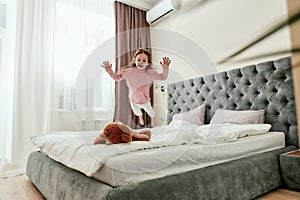 A cute small girl having fun and jumping down like a little tiger on her brown teddybear barefoot on a bed