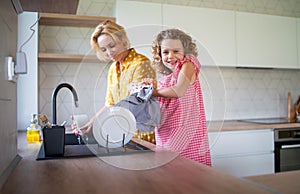 A cute small girl with mother indoors in kitchen at home, washing up dishes.