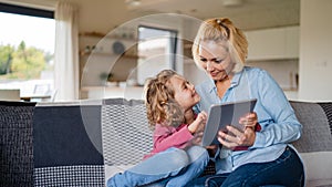 A cute small girl with mother on sofa indoors at home, using tablet.