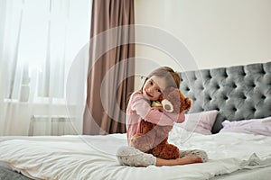 A cute small girl smiling and embracing a teddybear while sitting barefoot cross-legged on a bed at home