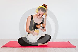 Cute small jack russell dog doing yoga on a mat at home with her owner. Young woman listening to music on yellow mobile phone and