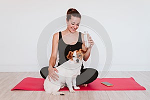 Cute small jack russell dog sitting on a yoga mat at home with her owner woman drinking water. Healthy lifestyle indoors