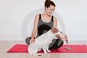 Cute small jack russell dog sitting on a yoga mat at home with her owner woman drinking water. Healthy lifestyle indoors