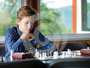 Cute, smart, 11 years old boy in shirt sits in the classroom and plays chess on the chessboard. Training, lesson, hobby, education