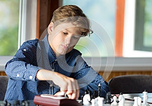 Cute, smart, 11 years old boy in shirt sits in the classroom and plays chess on the chessboard. Training, lesson, hobby, education