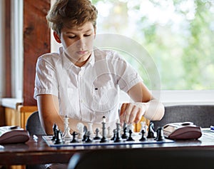 Cute, smart, 11 years old boy in white shirt sits in the classroom and plays chess on the chessboard. Training, lesson, hobby