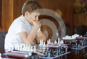 Cute, smart, 11 years old boy in white shirt sits in the classroom and plays chess on the chessboard. Training, lesson, hobby