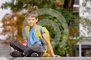 Cute smart boy with backpack sitting outdoors after school.