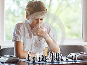 Cute, smart, 11 years old boy in white shirt sits in the classroom and plays chess on the chessboard. Training, lesson, hobby,