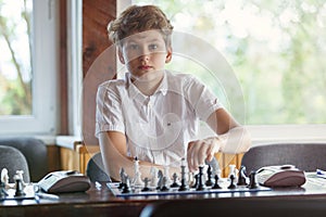 Cute, smart, 11 years old boy in white shirt sits in the classroom and plays chess on the chessboard. Training, lesson, hobby