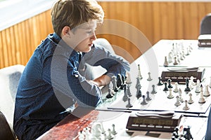 Cute, smart, 11 years old boy in white shirt sits in the classroom and plays chess on the chessboard. Training, lesson, hobby,