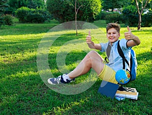 Cute, smart, young boy in blue shirt sits on the grass next to his school backpack, globe, chalkboard, workbooks. Education