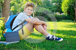 Cute, smart, young boy in blue shirt sits on the grass next to his school backpack, globe, chalkboard, workbooks. Education