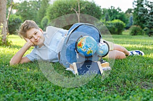 Cute, smart, young boy in blue shirt sits on the grass next to his school backpack, globe, chalkboard, workbooks. Education