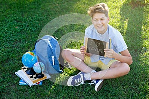 Cute, smart, young boy in blue shirt sits on the grass next to his school backpack, globe, chalkboard, workbooks. Education