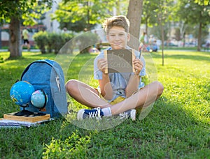 Cute, smart, young boy in blue shirt sits on the grass next to his school backpack, globe, chalkboard, workbooks. Education