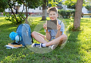 Cute, smart, young boy in blue shirt sits on the grass next to his school backpack, globe, chalkboard, workbooks. Education