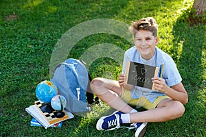 Cute, smart, young boy in blue shirt sits on the grass next to his school backpack, globe, chalkboard, workbooks. Education