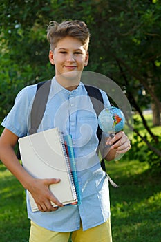 Cute, smart, young boy in blue shirt stands on the grass with globe and school backpack, workbooks. Education, back to school
