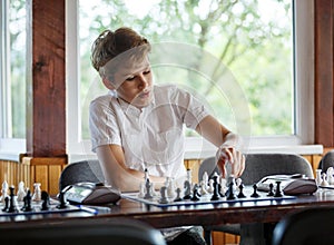 Cute, smart, young boy in white shirt plays chess on the chessboard in the classroom. Education, hobby, training