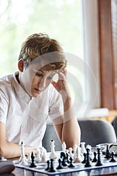 Cute, smart, young boy in white shirt plays chess on the chessboard in the classroom. Education, hobby, training