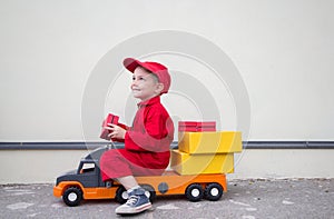 Cute smiling 3-4 year old boy in red overalls uniform and cap is sitting on big toy car truck with cardboard boxes