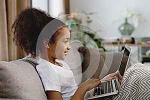 Cute smiling Afro-American little girl using laptop and smiling while sitting on sofa.