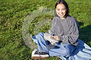Cute smiling asian girl sitting in park on blanket, reading and relaxing. Young beautiful woman enjoying sunny day with