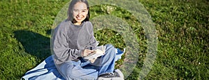 Cute smiling asian girl sitting in park on blanket, reading and relaxing. Young beautiful woman enjoying sunny day with