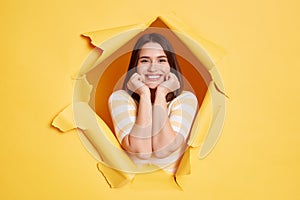 Cute smiling attractive woman stands in torn paper hole, looking through breakthrough of yellow background, looks at camera,