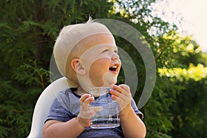 Cute smiling baby boy with a glass of water sitting in a baby chair against green bush