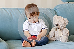 Cute smiling baby boy playing on smartphone sitting on sofa with teddy bear at home