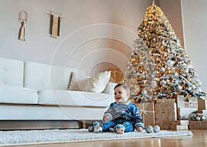 Cute smiling baby boy sitting on a white wool carpet in a bright cozy living room with a Christmas tree and many gift