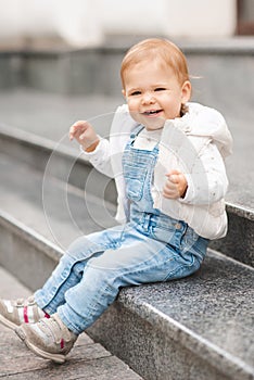 Cute smiling baby girl 1-2 year old wear casual denim pants and white top in city street outdoors. Looking at camera.