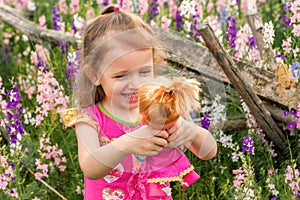 Cute smiling baby girl in bright dress plays with doll outdoors in green field