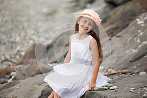 A cute smiling blonde girl 9 years old in a hat and white dress sits on the seashore