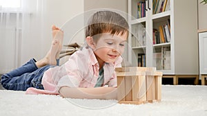 Cute smiling boy lying on floor and looking at toy house he built from toy wooden blocks. Concept of child education and