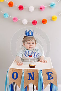 Cute smiling Caucasian baby boy in blue crown celebrating his first birthday at home. Child kid toddler sitting in high chair