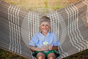 Cute smiling caucasian boy drinking water and having fun on hammock in backyard or outdoor. Kid swinging and relaxing in hammock