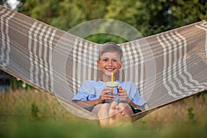 Cute smiling caucasian boy drinking water and having fun on hammock in backyard or outdoor. Kid swinging and relaxing in hammock