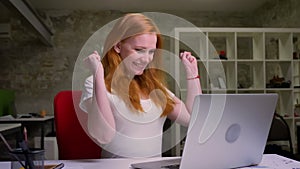 Cute smiling ginger caucasian girl is typing in her laptop while sitting in brick light office at desktop, natural face