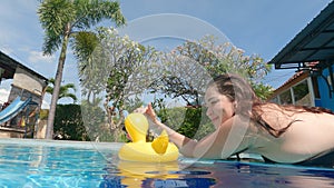 Cute smiling girl in bikini sunbathing and plays on poolside with yellow inflatable toy duck. Enjoy summer vacation in