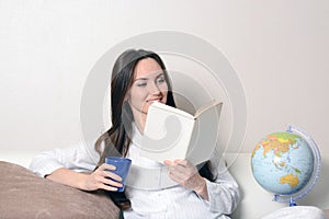 Cute smiling girl with a blue Cup and the diary, sitting on the couch in the room and reads.