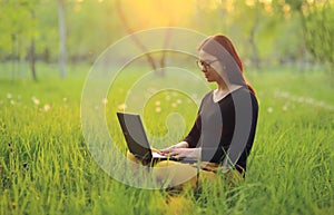 Cute smiling girl with laptop sits on green grass in the park and works remotely as a freelancer