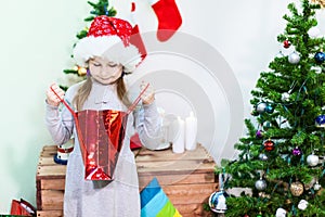 Cute smiling girl in red Santa hat looking inside of gift bag near Christmas tree, copyspace